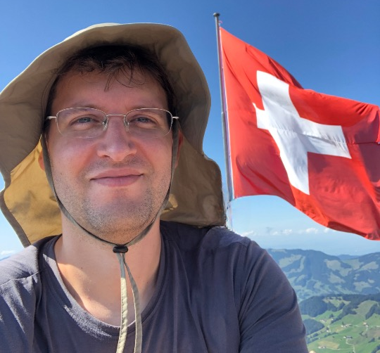 A profile picture of man wearing glasses and a hat. Behind him, there is a mountain landcape with a Swiss flag.
